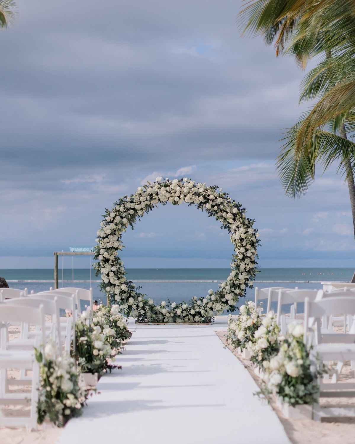 Round arch fully covered in flowers at the end of a beach aisle