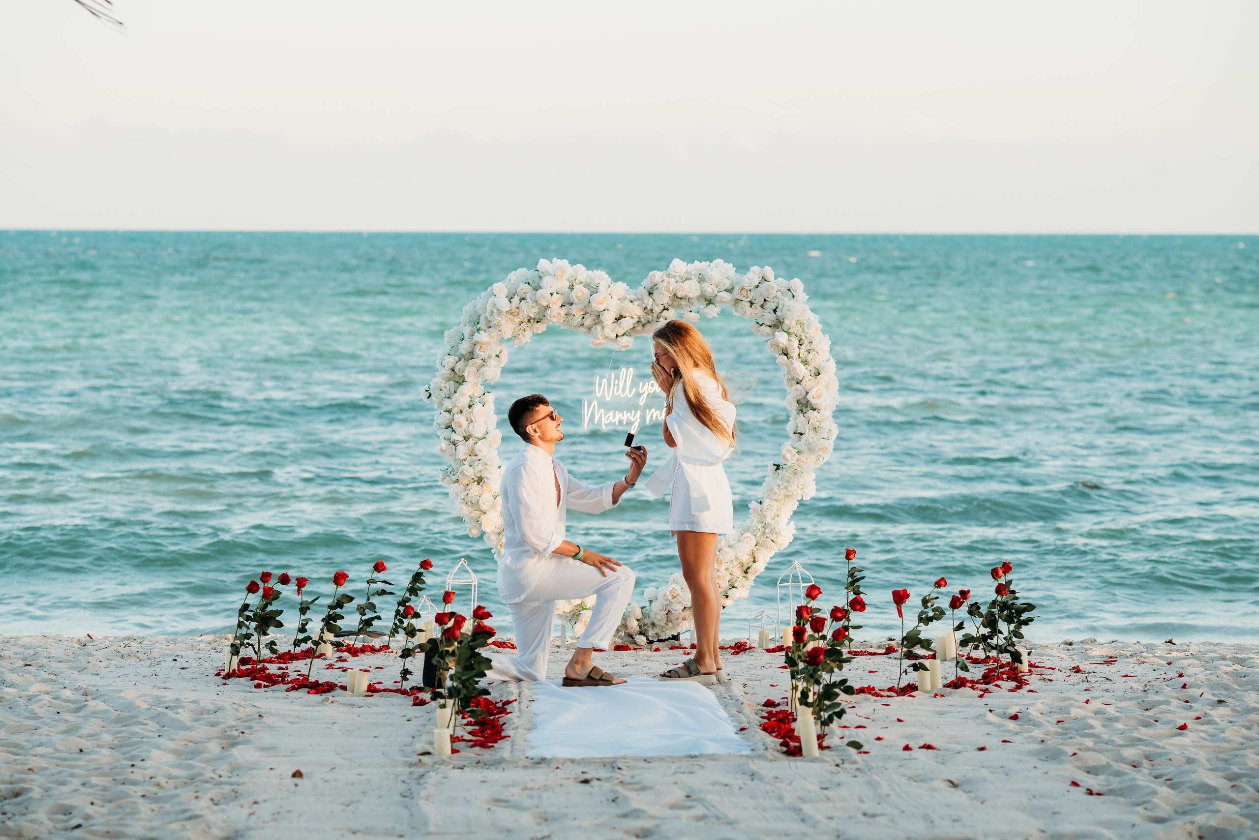 Man kneeling to propose under a white floral heart arch