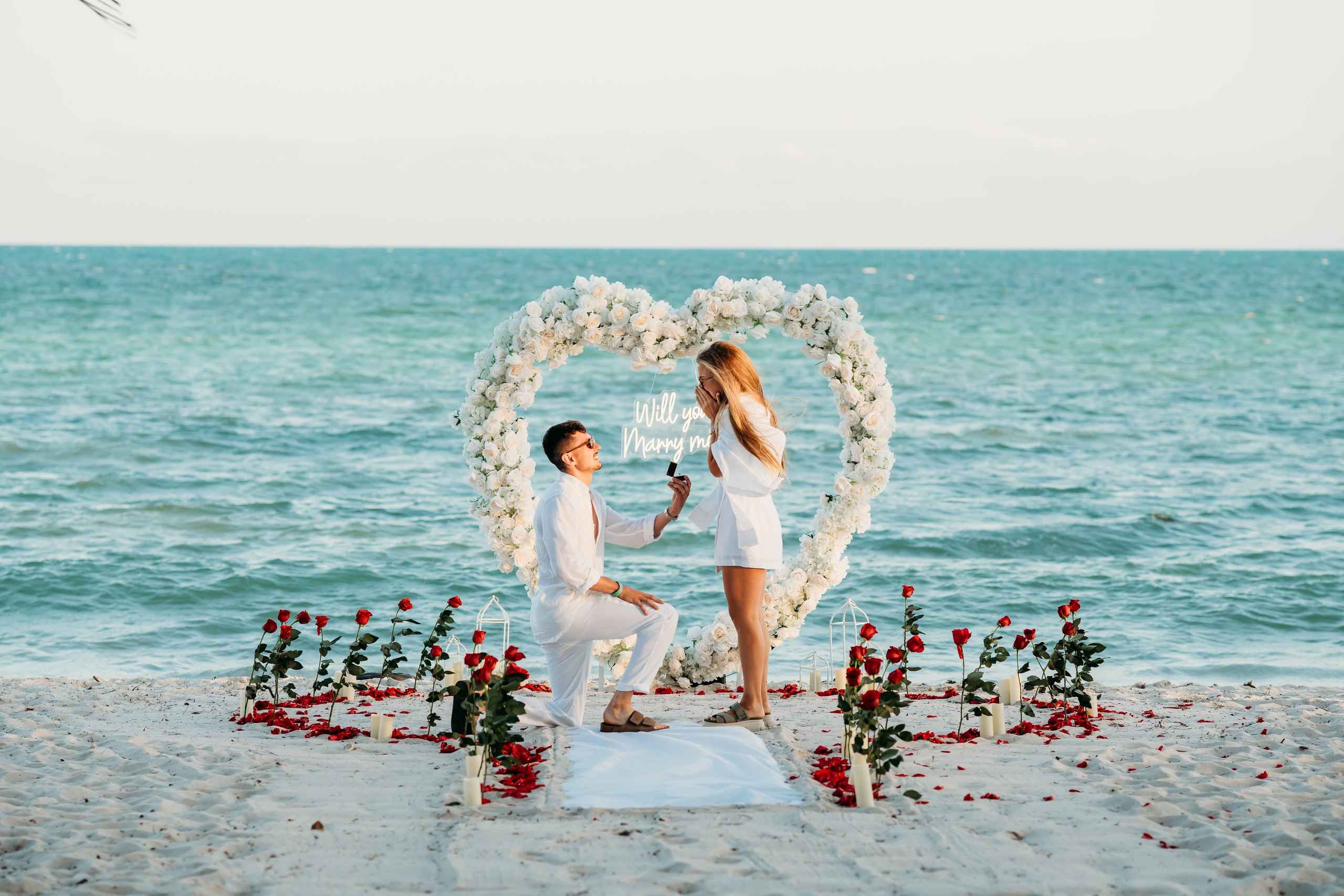 Man kneeling to propose under a white floral heart arch