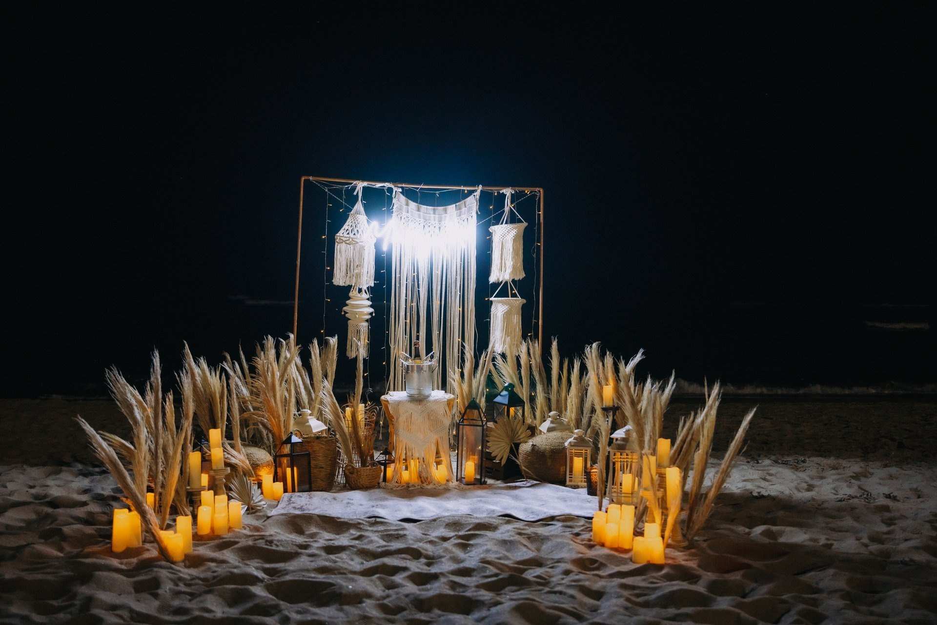 Macramé arch glowing at night on the beach, with pampas grass and lanterns in the sand